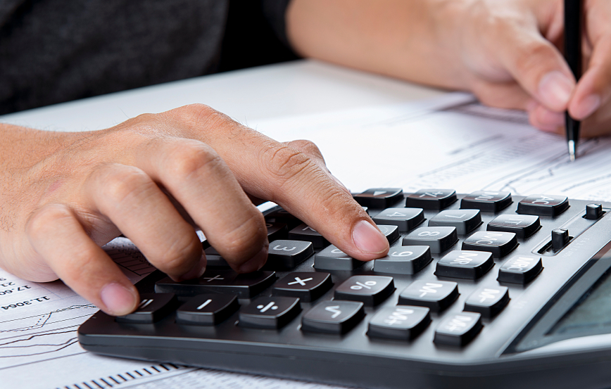 Photo of hands holding pen and pressing calculator buttons over documents