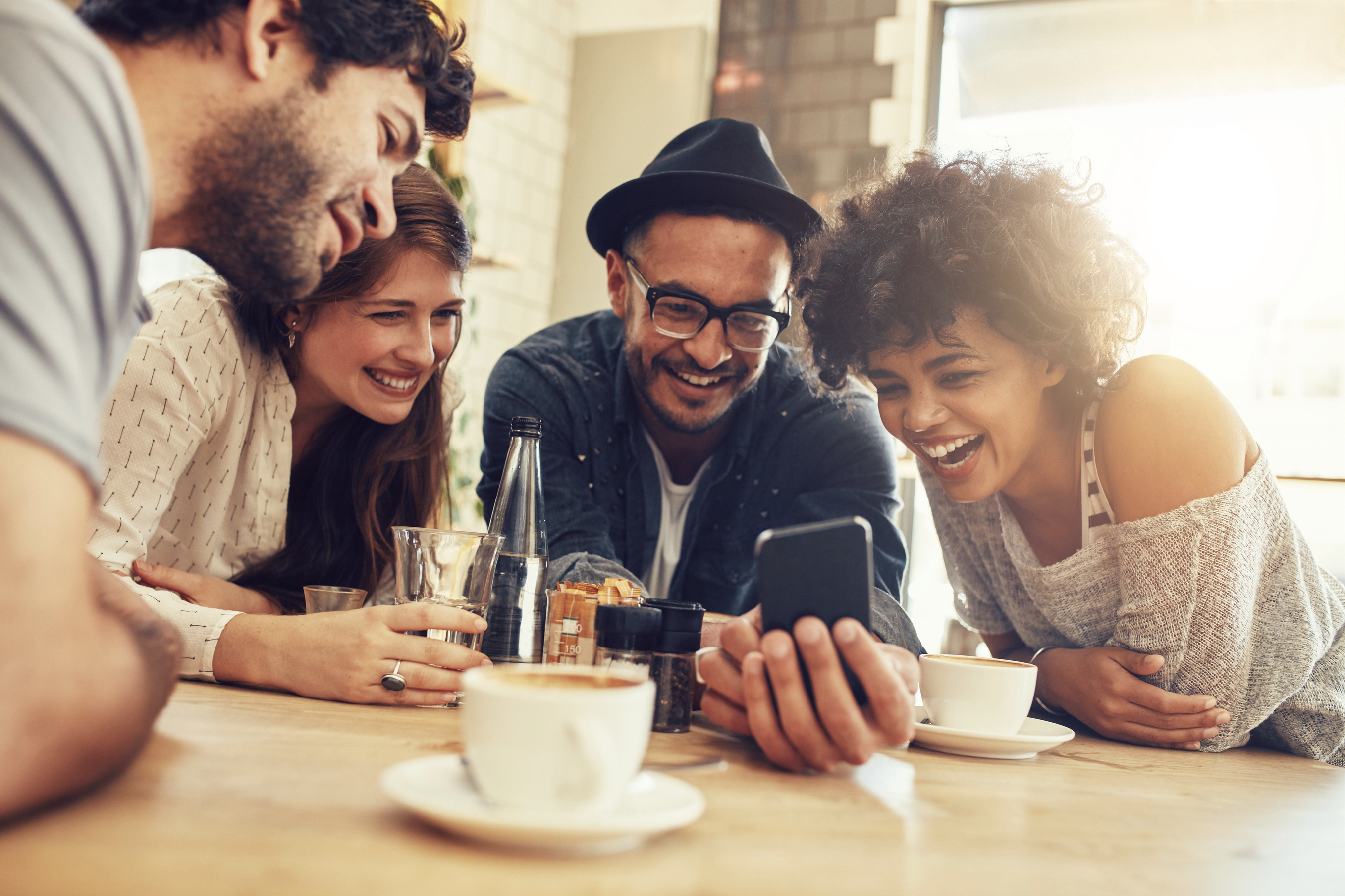 Portrait of cheerful young friends looking at smart phone while sitting in cafe. Mixed race people sitting at a table in restaurant using mobile phone.